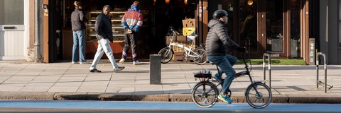 Photograph of Clapham High Street showing a shopfront and pavement scene. Several people are walking or standing outside the building, and a person on a bicycle is travelling along the street. The image captures the busy, everyday activity of Clapham High Street and is used to illustrate the location of a Local Plan consultation event.