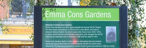Photograph of the entrance sign for Emma Cons Gardens. The green information board displays the name “Emma Cons Gardens” along with text about the history of the garden and a small portrait image. Trees, planting, and parts of surrounding buildings can be seen in the background.