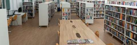 Photograph of the interior of South Lambeth Library. The image shows a bright library space with rows of bookshelves filled with books, wooden tables and chairs for reading and study, and high ceilings with large windows that allow natural light into the building. The library has a spacious, open-plan layout with shelving arranged in aisles and a central display stand.
