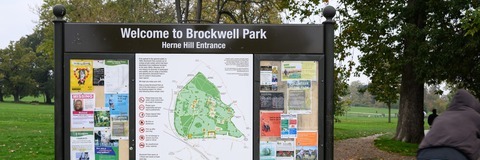 Information board displaying planning and development information in a public outdoor space. The board includes a large map at the centre, surrounded by notices, photographs, diagrams and text panels. The display is mounted within a dark timber-framed structure and is set against a green landscape with grass and trees in the background. The heading at the top of the board reads "Welcome to Brockwell Park".
