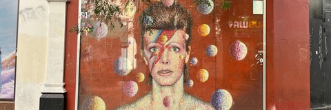 Large street mural on Tunstall Road in Brixton depicting David Bowie in his iconic Aladdin Sane lightning-bolt makeup. The colourful portrait is painted against a red background, surrounded by floating circular shapes, and is displayed behind a glass panel on the side of a building. Tree branches partially frame the mural above, with the pavement in the foreground.